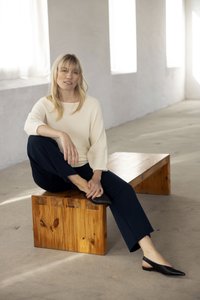 Femme aux cheveux blonds portant un pull crème et un pantalon noir assise sur un banc en bois dans une pièce lumineuse et minimalement meublée.