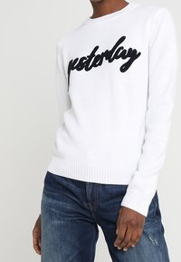 Biały sweter dzianinowy z okrągłym dekoltem i długimi rękawami. Na przodzie czarny napis „yesterday” wykonany z cekinów. Noszony z niebieskimi dżinsami.