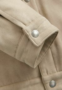 Mâneca unei jachete bej din suede cu un buton de prindere argintie. Materialul are un aspect moale și texturat, cu detalii de cusătură vizibile.