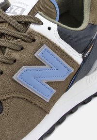 Pantof sport maro din piele întoarsă și plasă, cu logo albastru "N", șireturi groase maro, talpă mijlocie albă și talpă exterioară neagră cu model texturat.