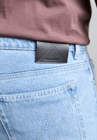 Lys blå denimjeans med et svart lærmerke som har et diamantmønster og "Nanushka" preget, plassert mot en brun topp.