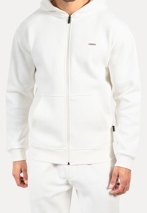 Mand iført en simpel hvid zip-up hættetrøje med forlommer og matchende hvide bukser, stående mod en hvid baggrund.