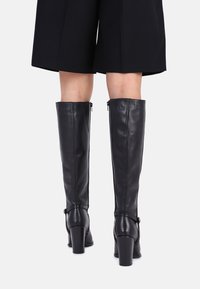 Bottes noires en cuir montant jusqu'aux genoux avec talons épais, fermetures éclair latérales et sangles décoratives à la cheville. Texture lisse et design élégant.