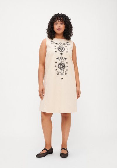Robe sans manches beige avec broderie florale noire sur le devant. Tissu lisse avec une coupe décontractée, associée à des chaussures en mesh noires.