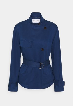 Marineblaue Jacke mit Gürtel und Stehkragen, drei schwarzen Knöpfen und drei Klappentaschen, dargestellt vor weißem Hintergrund.