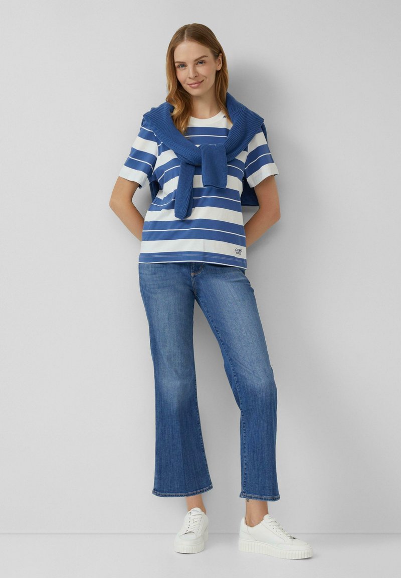 Blauw-wit gestreept shirt met korte mouwen, met een blauwe gebreide trui over de schouders gedrapeerd, gecombineerd met lichtblauwe flared jeans en witte sneakers.