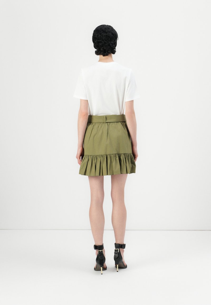 Gonna verde oliva a balze con vita con cintura, abbinata a una camicia bianca a maniche corte e tacchi neri con cinturino alla caviglia.