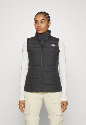Chaleco aislante negro con cuello alto, cierre de cremallera y costuras horizontales. Lleva puesto sobre una camiseta de manga larga blanca, con el logo en el pecho.