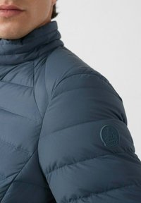 Gevoerde jas in donkerblauw met een hoge kraag, voorzien van horizontale quiltlijnen en een rubberen logo-patch op de linker mouw.