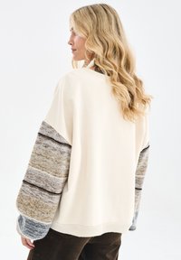 Kremowy sweter z teksturowanymi, wielokolorowymi rękawami w paski w odcieniach szarości i beżu. Luźny krój, ściągacze na mankietach i przy dekolcie.