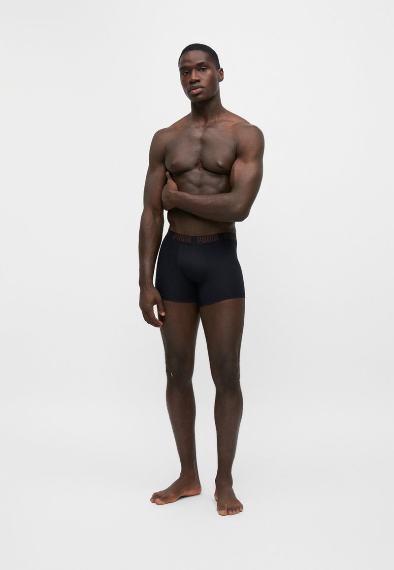Homme musclé debout pieds nus portant un boxer noir Puma sur un fond blanc uni, avec un bras croisé sur la poitrine.