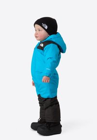 Blå barnedress med svarte aksenter, hette og elastiske mansjetter; har vanntett materiale og isolerte svarte snøstøvler.