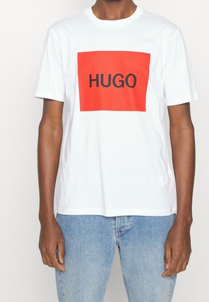 White cotton t-shirt featuring a large red rectangular graphic with the word "HUGO" in bold black font. Short sleeves and a crewneck design.