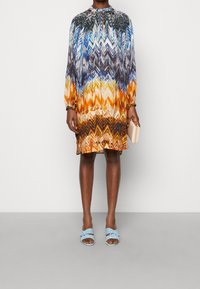 Flerfärgad chevronmönstrad klänning med långa ärmar och hög krage. Bärs med ljusblå sandaler och håller en liten beige kuvertväska.