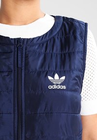 Navyblå pufferväst med dragkedja fram, horisontellt quiltad textur och Adidas-logotyp i vitt på den vänstra bröstområdet.