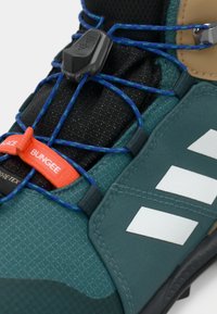 Shoe de caminhada verde e preto com material GORE-TEX, apresentando cordões elásticos azuis, um botão laranja e detalhes refletivos em prata.