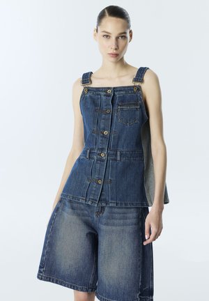 Kobieta ubrana w bezrękawnikowy, zapinany na guziki denimowy top z przednią kieszonką na piersi i luźne denimowe szorty, stoi na tle jednolitego jasnego tła.