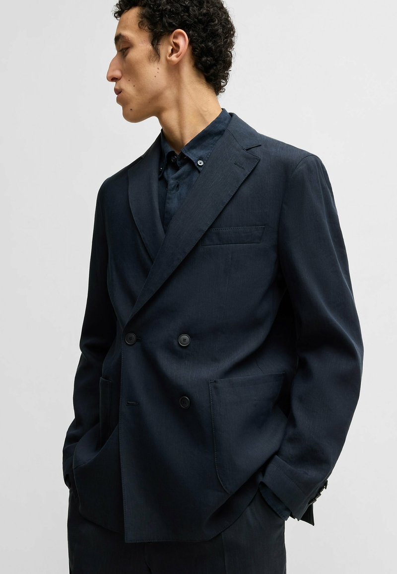 Marineblaues Doppelreiher-Blazer aus strukturiertem Stoff mit spitzen Revers und zwei Fronttaschen, getragen über einem dunklen Hemd.