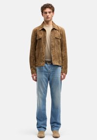 Giacca in camoscio marrone con bottoni sul davanti, abbinata a una camicia beige e jeans azzurri. Tessuto liscio e vestibilità classica.