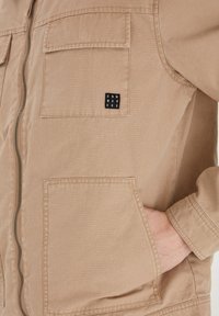 Beige jakke med glidelås foran, to brystlommer og et logo-patch. Laget av et teksturert stoff, med lange ermer og en avslappet passform.