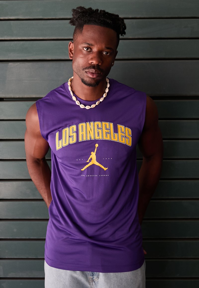 Tricou fără mâneci de culoare violetă, cu textul "LOS ANGELES" în galben și un logo galben în formă de siluetă pe față, asociat cu pantaloni scurți din denim deschis.