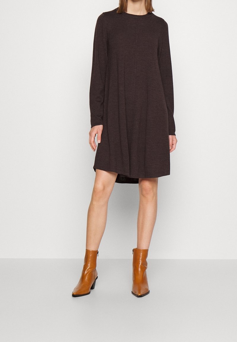 Femme portant une robe longue marron foncé à manches longues jusqu'aux genoux, associée à des bottines en cuir marron clair, debout devant un fond clair uni.