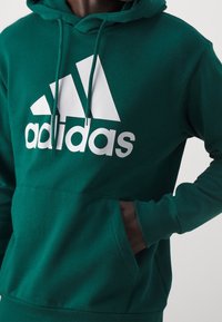 Sudadera con capucha verde, hecha de tela suave, que presenta un gran logotipo blanco de Adidas y un bolsillo tipo canguro en la parte delantera.