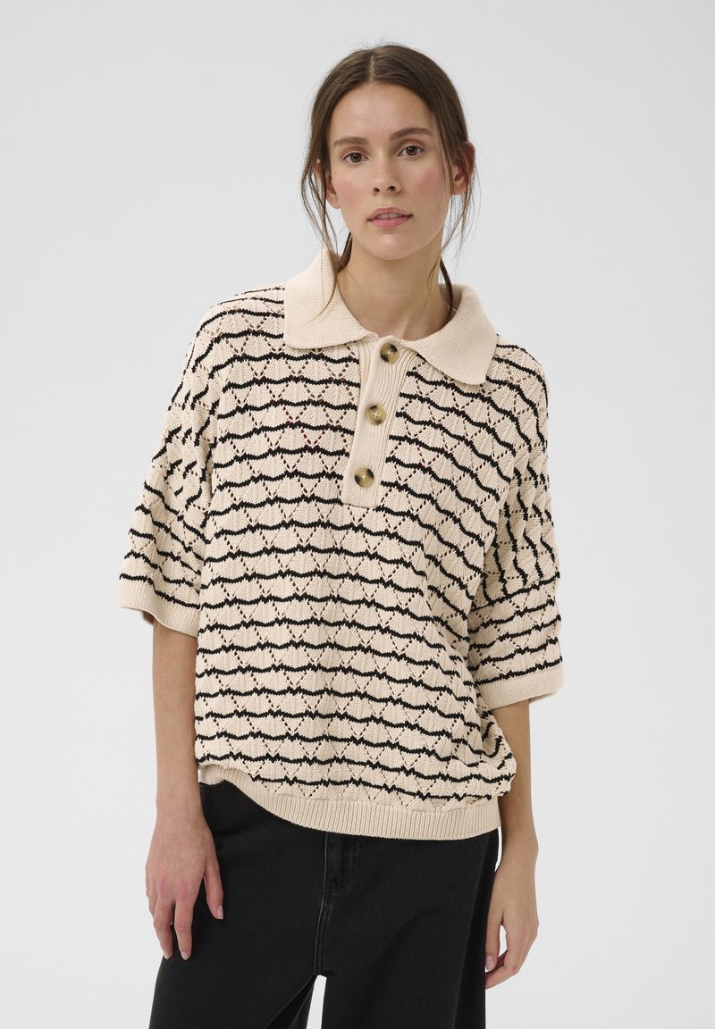 Femme portant un pull polo en maille beige avec un motif noir en forme de coquille et trois grands boutons, associée à un pantalon noir, debout devant un fond uni.