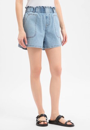 Vrouw draagt lichtblauwe denimshort met elastische tailleband, voorzakken en zwarte sandalen met gesp.