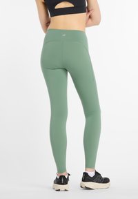 Leggings en verde apagado, textura suave, cintura alta, bolsillo lateral, combinados con zapatillas deportivas negras, con suela texturizada y logotipo.