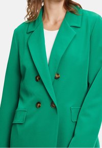 Mujer con un blazer verde brillante de doble botonadura con botones de carey y bolsillos con solapa sobre una blusa blanca.