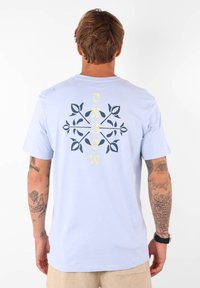 T-shirt bleu clair avec un graphique central en bleu foncé et jaune comportant des motifs de feuilles et le mot "Oxbow". Fabriqué à partir d'un tissu à la texture lisse.