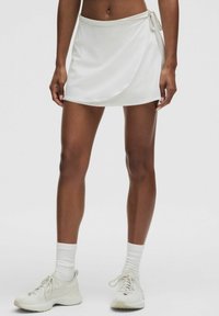 Skort atlético blanco con diseño envolvente, fabricado en una tela suave. Presenta un lazo en el lado y se combina con zapatillas y calcetines de color claro.