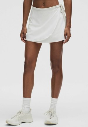 White athletic skort with a wrap design, made from smooth fabric. Features a tie on the side and is paired with light-colored sneakers and socks.