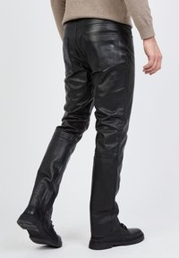 Schwarze Lederhosen mit engem Schnitt, glatter Textur und Gesäßtaschen. Kombiniert mit flachen schwarzen Schuhen.