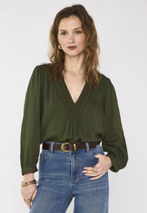 Femme aux cheveux bruns ondulés portant un chemisier vert olive à col en V, un jean bleu, une ceinture marron, des boucles d'oreilles dorées, des bracelets et des bagues.