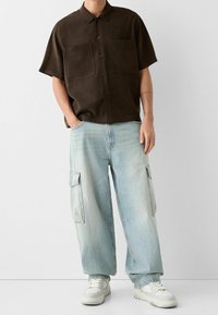 Mężczyzna w ciemnobrązowej koszuli z krótkim rękawem zapinanej na guziki, jasnoniebieskich szerokich dżinsach cargo oraz białych trampkach, stoi z jedną ręką w kieszeni.