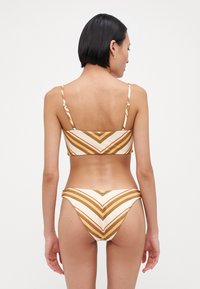 Bikini set z belo osnovo in zlate in oranžne črte v obliko chevron. Material se zdi teksturiran, z nastavljivimi naramnicami in minimalistično opremo.