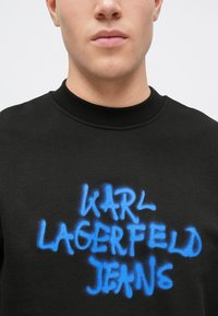 Svart hettegenser med rund halsutringning som har dristig, blå graffiti-tekst som sier "KARL LAGERFELD JEANS." Myk, teksturert stoff.