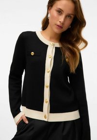 Donna che indossa un cardigan nero con rifiniture crema e bottoni dorati, mani nelle tasche dei pantaloni neri, lunghi capelli mossi e orecchini d'argento.
