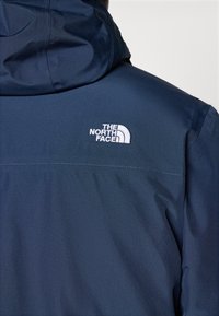 Veste bleu foncé avec capuche, montrant le logo blanc « The North Face » brodé sur le haut du dos.