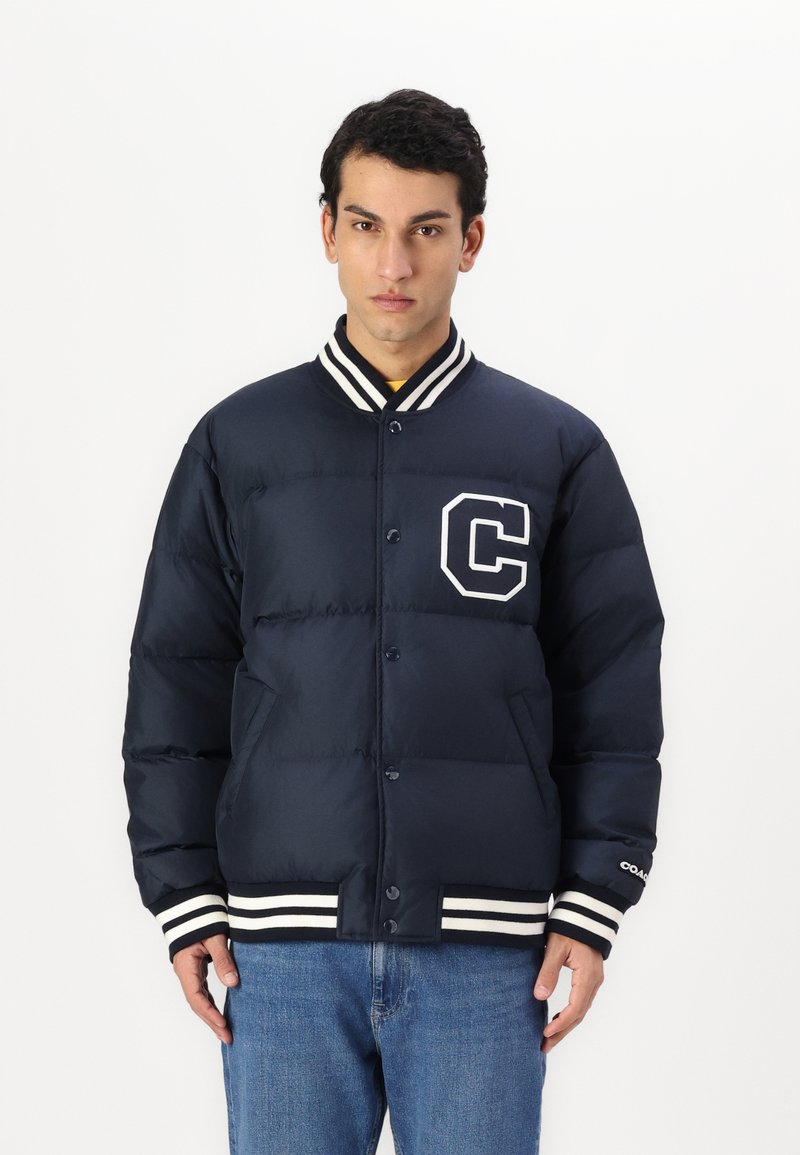 Námornícky modrá prešívaná bunda s bielym pruhovaným žebrovaným golierom a manžetami, s bielym logom "C" na ľavej strane hrudi a s gombíkovým zapínaním.