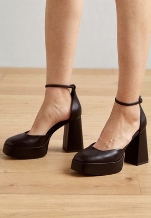 Femme portant des chaussures noires à plateforme à talons hauts avec des brides à la cheville sur un sol en bois.