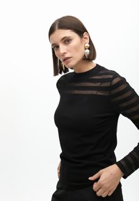 Čierny top s dlhým rukávom, ktorý má priesvitné pruhované detaily na rukávoch, okrúhly výstrih a priliehavý strih. Obsahuje malú vyšívanú ozdobu na hrudi.