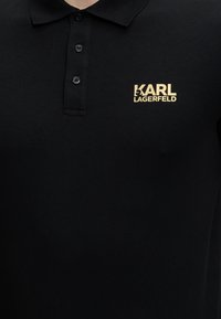 Μαύρο πόλο με τρία κουμπιά και κίτρινο λογότυπο "KARL LAGERFELD" στο αριστερό μέρος του στήθους.