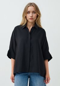 Black short-sleeve button-up shirt in a lightweight fabric with a relaxed fit and folded cuffs, paired with blue straight-leg jeans.