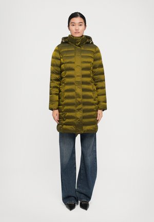 Vrouw in een olijfgroen gewatteerd lange pufferjack met capuchon, brede blauwe jeans en zwarte spitse schoenen, staand tegen een effen achtergrond.