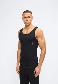 Άνδρας με κοντά μαλλιά, που φοράει στενή μαύρη φανέλα με κόκκινο λογότυπο "HUGO", στέκεται μπροστά σε απλό λευκό φόντο.