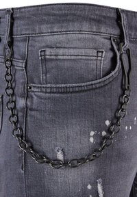 Šedé džíny z denimu s distorzovanými časťami a reťazovým doplnkom pripevneným na zadnej strane. Obsahuje detailný pohľad na vrecko a pás.