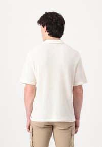 Hvit, teksturert piqué-skjorte med krage og korte ermer, iført med beige cargo-shorts, vist fra baksiden.